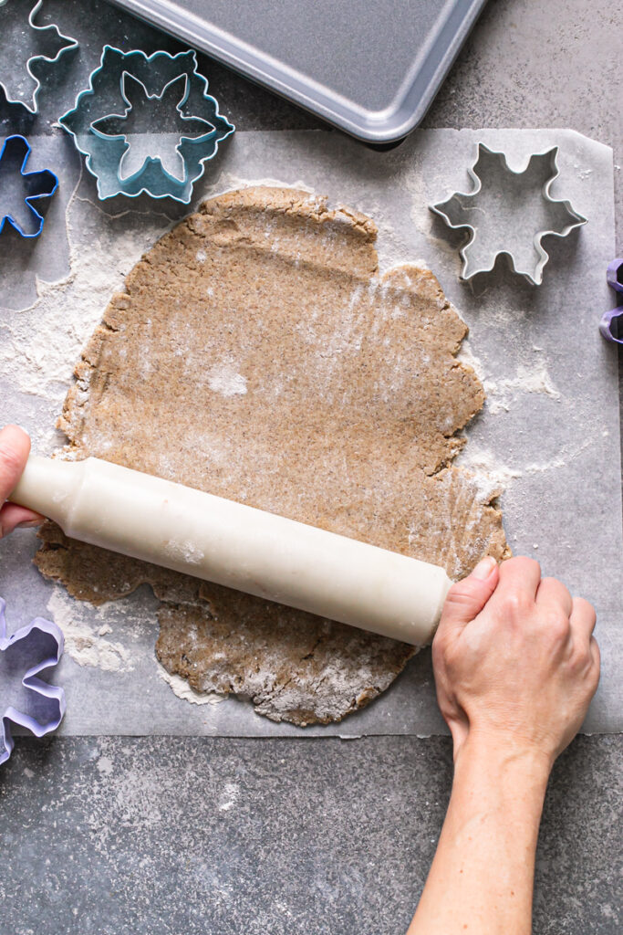 action shot of rolling the dough out with a rolling pin on top of a piece of parchment paper and the snowflake cookie cutters all around it