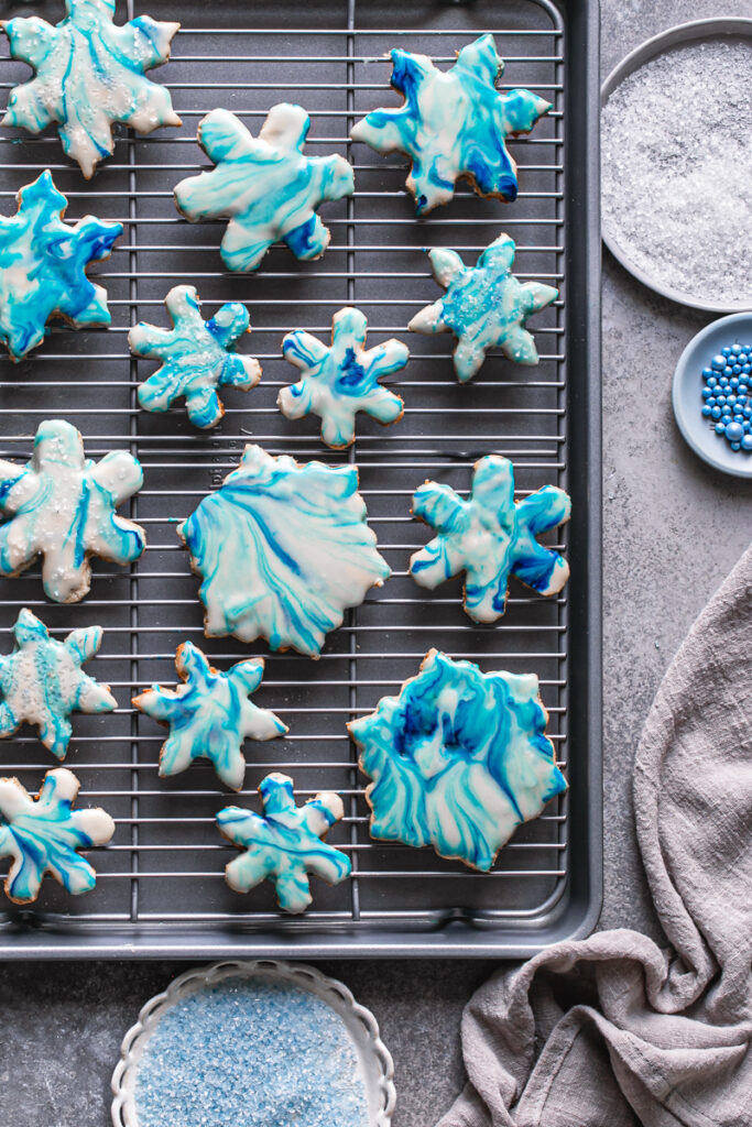 decorated grain-free marbled snowflake sugar cookies on a wire baking rack on top of a baking sheet with the sparkling sugar and candy pearls beside it