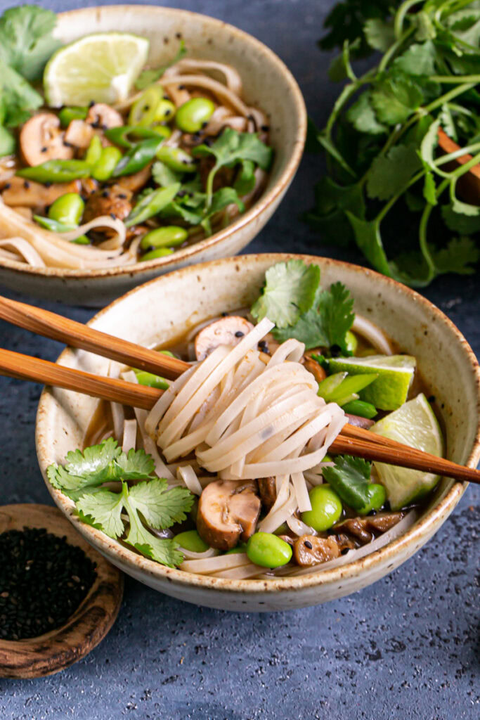 3/4 shot of two bowls of mushroom pho, the front bowl with noodles wrapped around the chopsticks out of the bowl