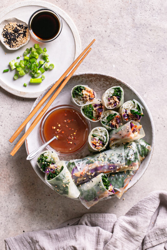 a bowl of sesame kale spring rolls, some cut in half to reveal the filling, a plate with soy sauce, sliced green onion and sesame seeds next to the bowl and a linen napkin