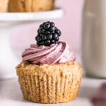 a single vanilla frosted cupcake with a blackberry on top, a jar of almond milk in the background and a cake stand of more cupcakes in the background