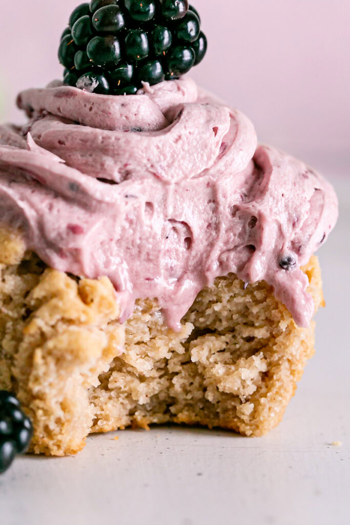 a close up shot of a vanilla cupcake frosted with blackberry frosting with a bite taken out of it