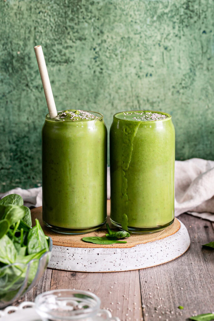 two glasses of Anti-Inflammatory Vegan Green Smoothie next to each other on an upside down plate with a bowl of spinach beside it
