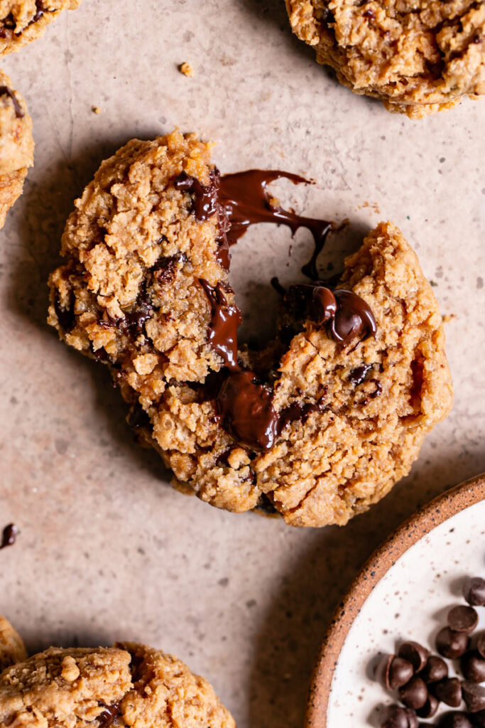 a Quinoa Chocolate Chip Cookie broken with melted chocolate oozing from the middle and a plate of chocolate chips in the corner with other cookies around it