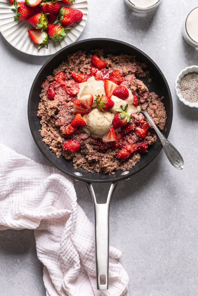 a skillet of strawberries and cream breakfast quinoa topped with fresh strawberries and banana nice cream sprinkled with chia seeds. Two glasses of almond milk and a plate of fresh strawberries above it