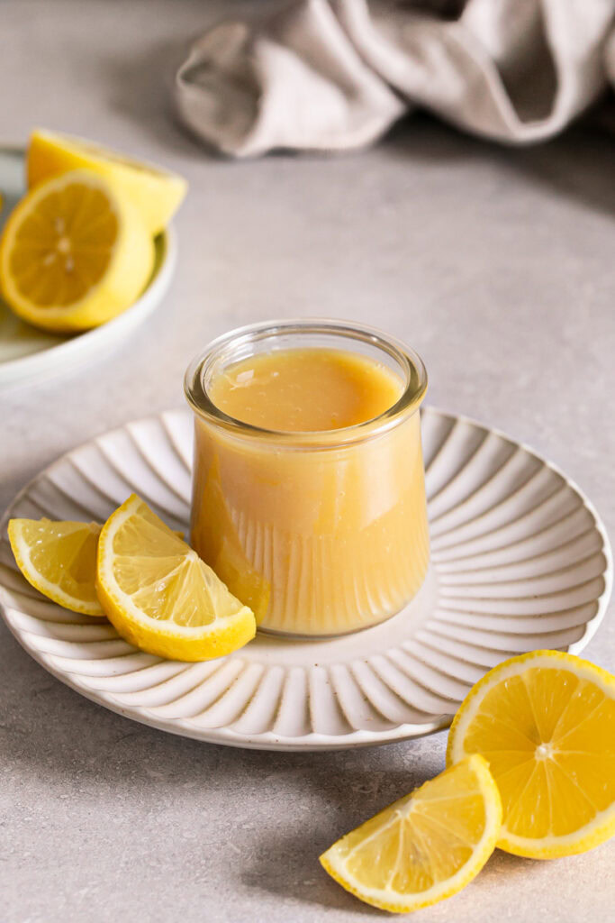a jar of lemon curd on a plate with fresh lemons around it.