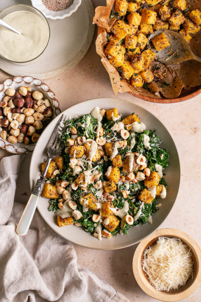 Kale Caesar Salad with Polenta Croutons and Toasted Hazelnuts, caesar dressing on the side with a bowl of vegan parmesan cheese, toasted hazelnuts and polenta croutons beside the salad