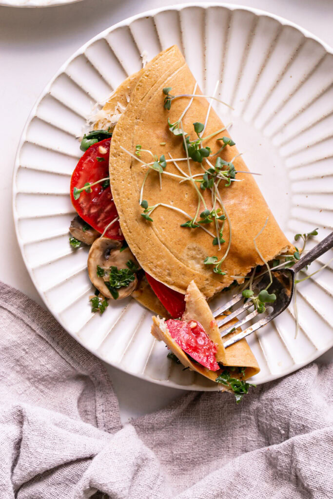 Vegan Gluten-Free Savory Pancake filled with vegan cheese, mushroom, kale and onion, and tomato on a plate garnished with fresh microgreens a bite on a fork