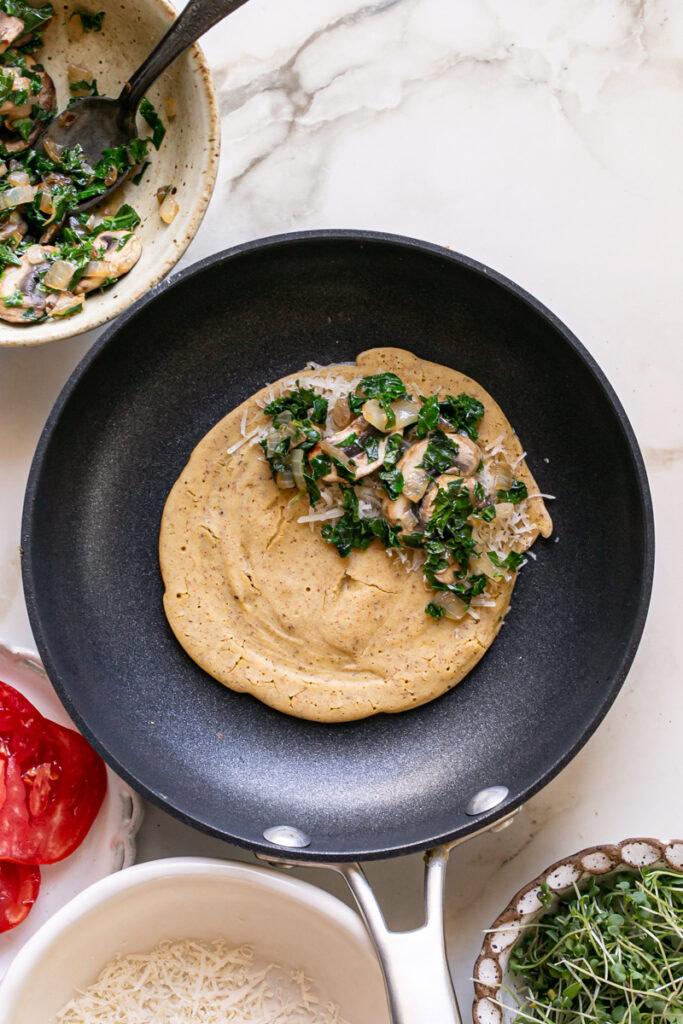 vegan gluten-free savory pancake in a skillet with vegan cheese, sautéed kale, mushroom, and onion on half of the pancake