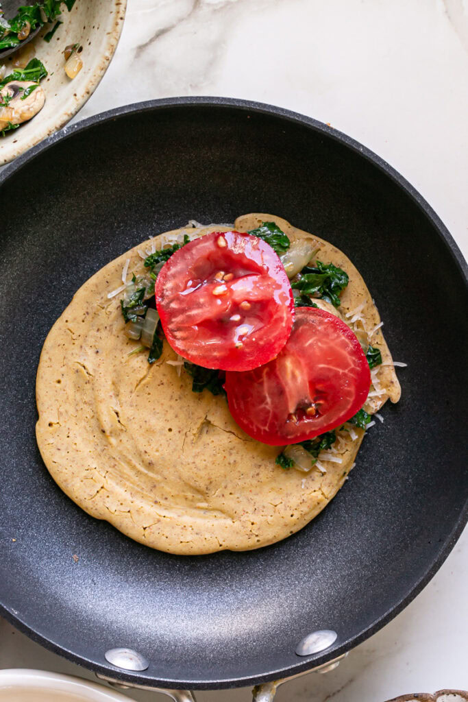 vegan gluten-free savory pancake in a skillet with vegan cheese, sautéed kale, mushroom, and onion, and fresh tomato on half of the pancake