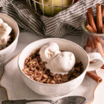 a close up of two mugs of Cinnamon Apple Crisp in a Mug topped with vegan vanilla ice cream, a basket of apples in the background and a jar of cinnamon sticks