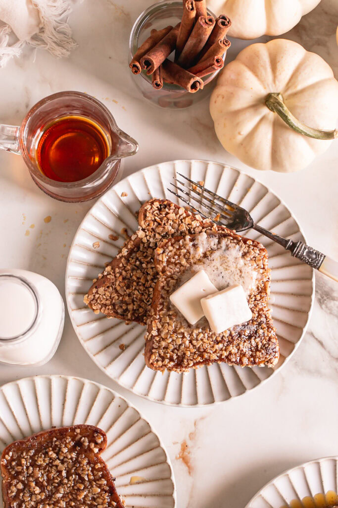 two slices of pumpkin spice oat crusted French toast on a plate topped with two squares of butter, a fork on the plate, a jug of plant milk beside it, a glass with maple syrup above the plate, a white pumpkin and a glass of cinnamon sticks above it