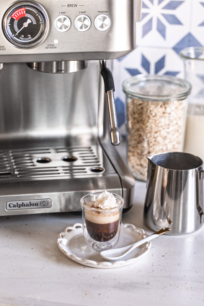 a small cup of apple crisp oat milk macchiato with the calphalon espresso machine in the background along with a jar of oats, a jug of oat milk, and a steel milk jug