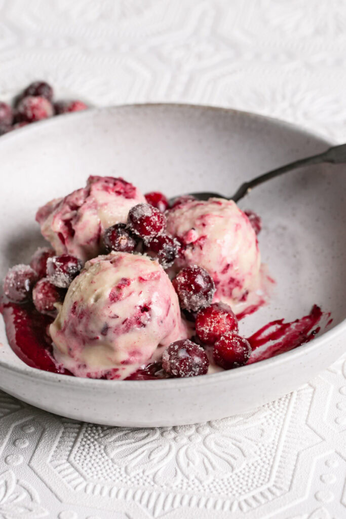 3/4 shot of three scoops of homemade dairy free cranberry swirl ice cream in a bowl with a spoon topped with sugared cranberries