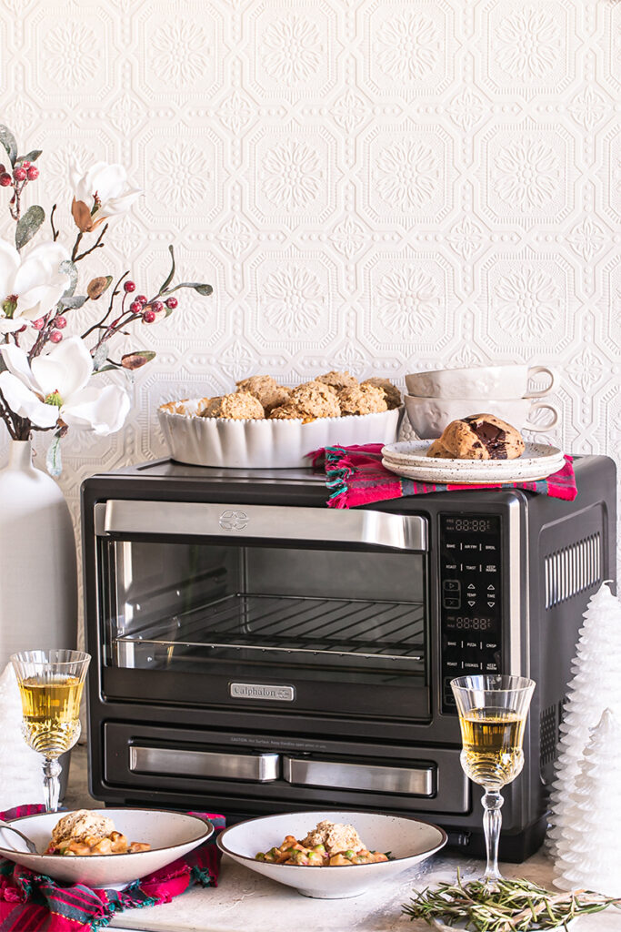 a prepared vegan dinner served in bowls sitting in front of Calphlaon Air fry oven with two glasses of wine next to it, holiday decor, and two chocolate chunk cookies on top of the oven with two mugs stacked inside one another