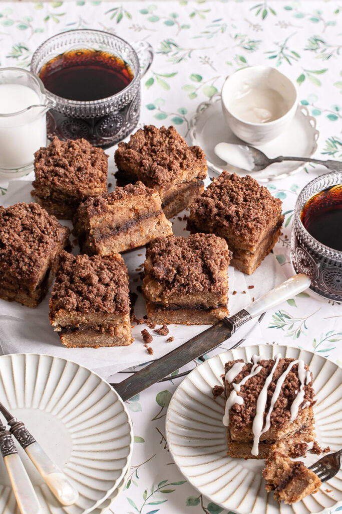 almond flour coffee cake sliced into squares on a parchment lined wire cooking rack with two mugs of coffee, a stack of plates, a bowl of icing, creamer, and an iced coffee cake square on a plate with a fork