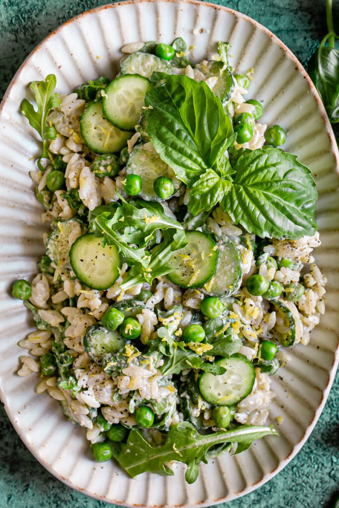 a close up of lemon basil orzo salad with peas, cucumber, and arugula on an oval plate garnished with fresh basil