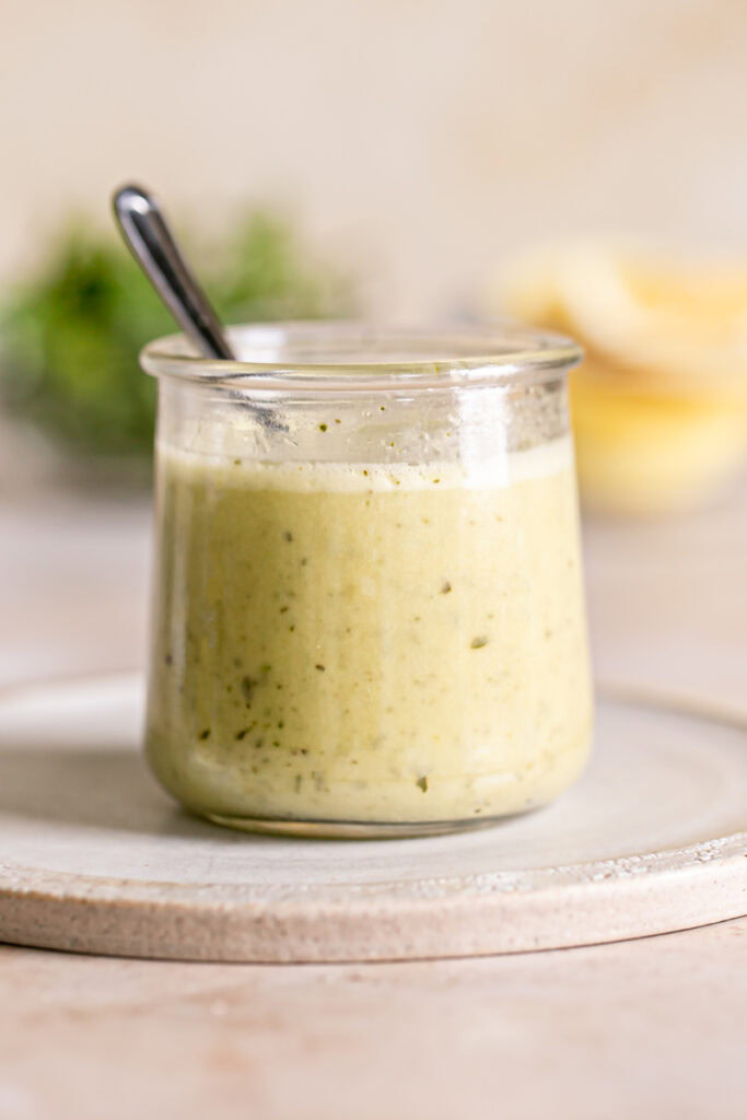 a jar of Lemon Basil Vinaigrette Dressing on a plate with a spoon in it, fresh basil leaves and lemon blurred in the background