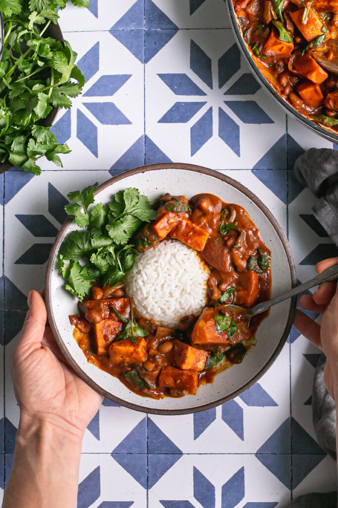 hands holding a bowl of sweet potato black eyed pea curry, scooping some out on the spoon ready to eat it, a plate of fresh cilantro above it