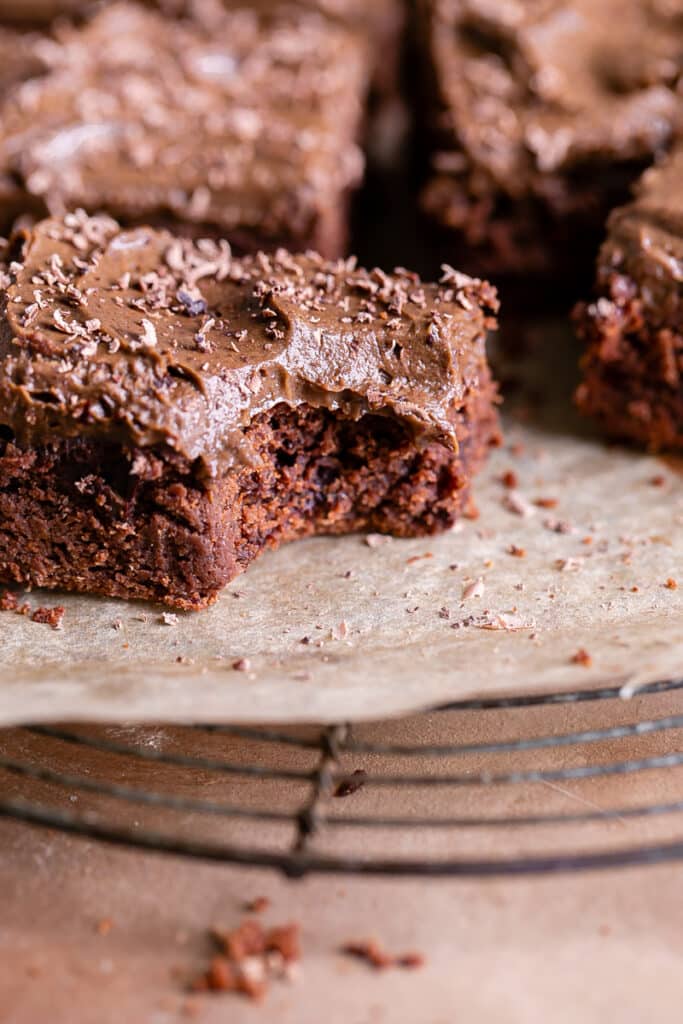 a close up of butternut squash brownies on a parchment lined wire cooling rack topped with chocoalte avocado frosting, one brownie with a bite taken out of it