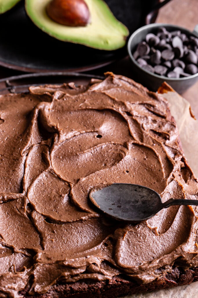 action shot of swirling the avocado chocolate frosting with a spoon on brownies with an avocado and a cup of dark chocolate chips in the background