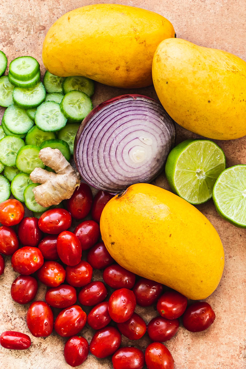 sliced mini cucumbers, cherry grape tomatoes, half a red onion, 3 mangos, a lime cut in half, and a whole piece of ginger