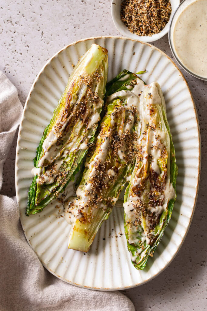three halves of romaine lettuce heads on an oval plate drizzled with caesar dressing and sprinkled with gluten-free toasted breadcrumbs. A bowl of bread crumbs and caesar dressing above the oval plate.