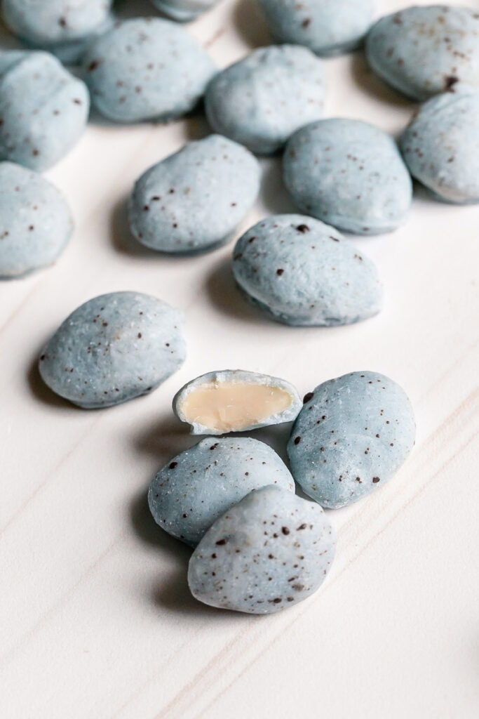 a pile of white chocolate covered almonds colored robin's egg blue using blue spirulina and speckled with dark chocolate one almond cut in half to reveal the almond inside.