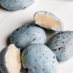 a close up of a pile of white chocolate covered almonds colored robin's egg blue using blue spirulina and speckled with dark chocolate with two almonds cut in half to reveal the almond inside.