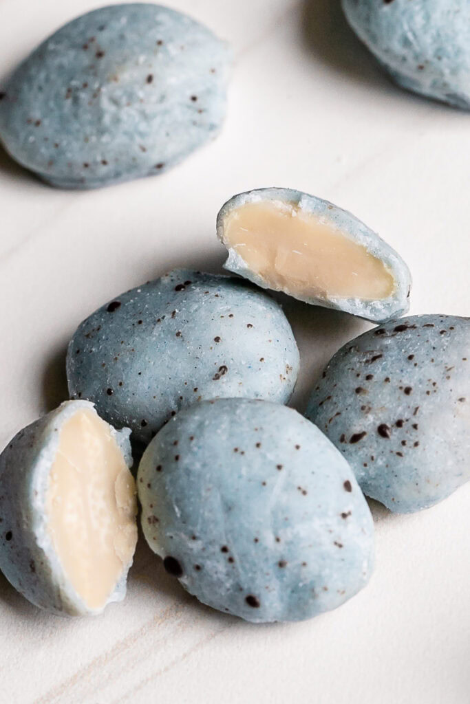 a close up of a pile of white chocolate covered almonds colored robin's egg blue using blue spirulina and speckled with dark chocolate with two almonds cut in half to reveal the almond inside