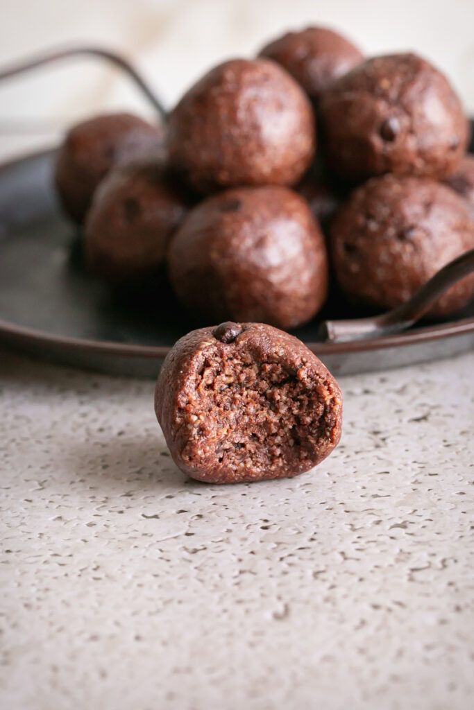 a single sunflower seed chocolate brownie bit on a countertop with a bite taken out of it and the other rolled brownie bites stacked on a plate behind it
