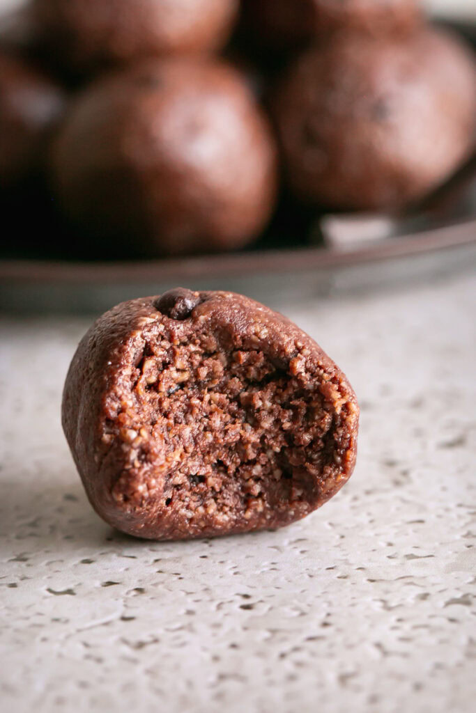a close up shot of a single sunflower seed chocolate brownie bit on a countertop with a bite taken out of it and the other rolled brownie bites stacked on a plate behind it