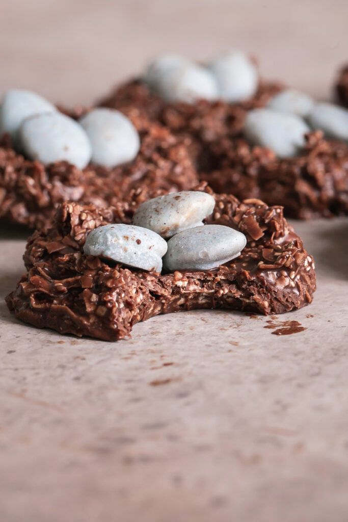 3/4 shot of a Toasted Coconut Chocolate Bird's Egg Nest Cookie filled with three robin's egg white chocolate covered almonds in it and a bite taken out of the cookie with other cookies in the background