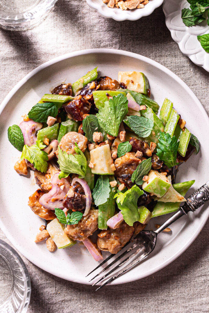 close up of a plate of Ants On A Log Celery date salad with plant-based Chicken, a fork on the plate, a bowl or crushed roasted peanuts, a small plate of fresh mint, and two glasses of water on a linen tablecloth