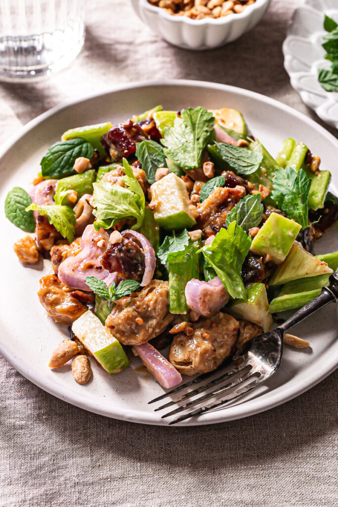 3/4 shot of a plate of Ants On A Log Celery date salad with plant-based Chicken, a fork on the plate, a bowl or crushed roasted peanuts, a small plate of fresh mint, and two glasses of water on a linen tablecloth
