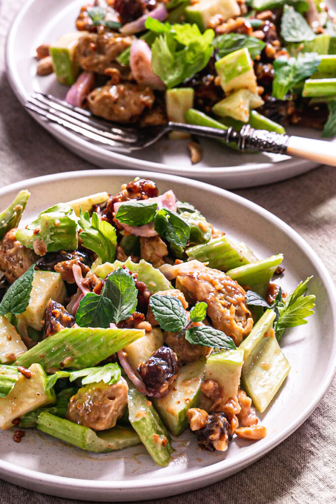 close up 3/4 image of two plates of a plate of Ants On A Log Celery date salad with plant-based Chicken, a fork on the plate on a linen tablecloth