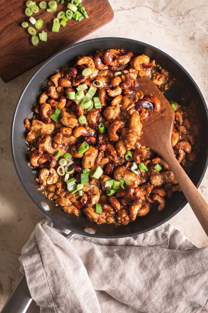 cooked vegan sticky cashew chicken in a skillet garnished with sliced green onions and a wooden spatula in the pan