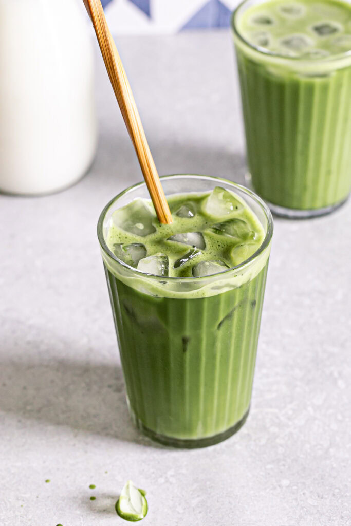 two glasses of iced matcha lattes, one glass with a wooden stir stick sticking out of it and a jug of rice milk in the background