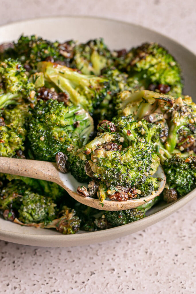 3/4 image of a spoon scooping broccoli tossed with fresh dill, raisins, and harissa tahini dressing out of a bowl