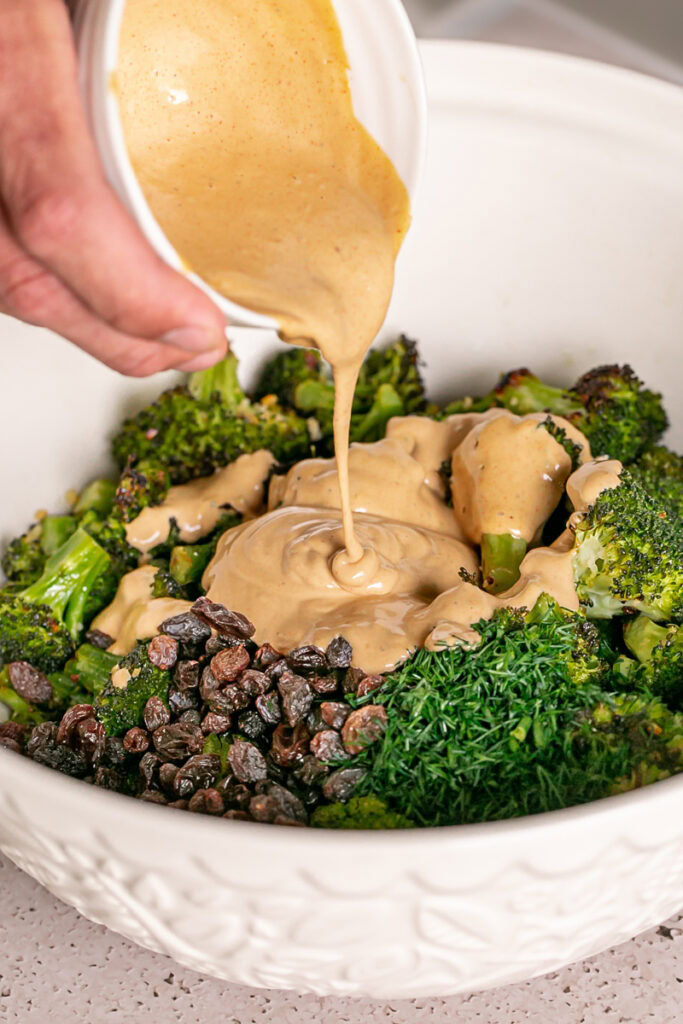 pouring the harissa tahini sauce over the broccoli in a large bowl with the fresh dill and raisins