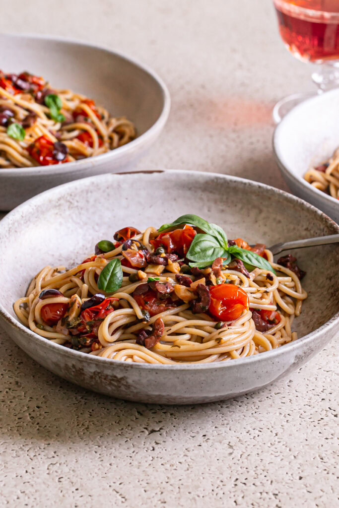 3/4 shot of 3 bowls of Cherry Tomato Puttanesca spaghetti with a glass of wine in the background
