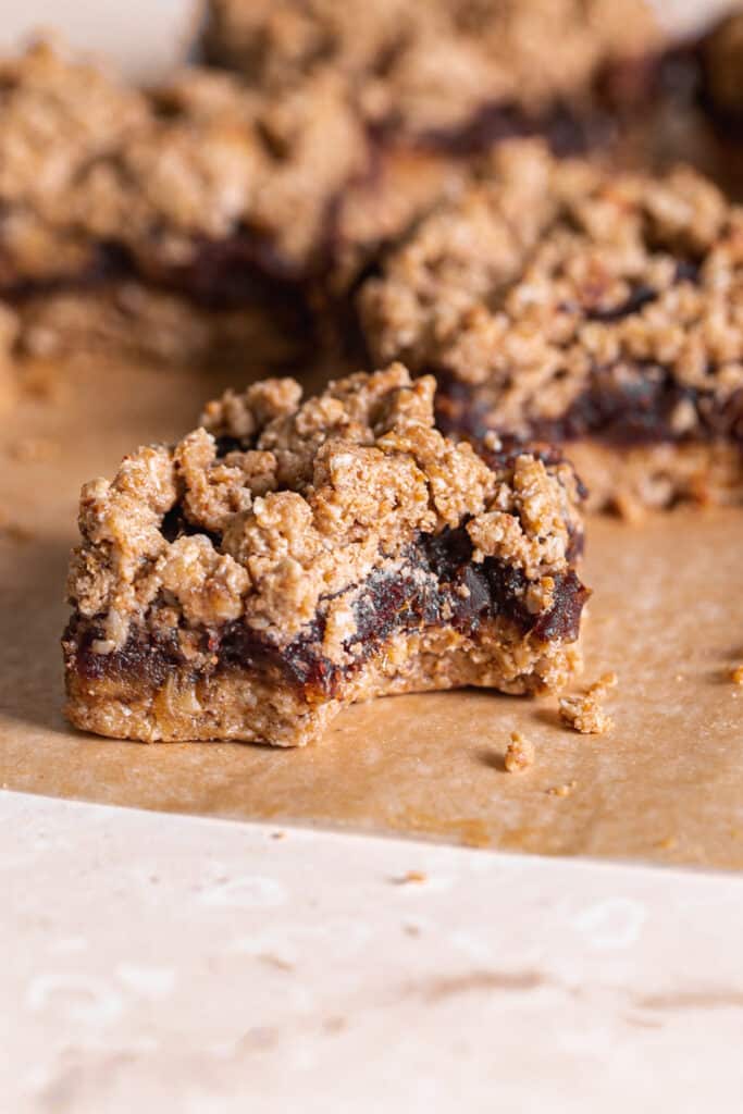 a single Peanut Butter Oat Date Crumb Bar with a bite taken out of it on a piece of parchment paper with more bars behind it