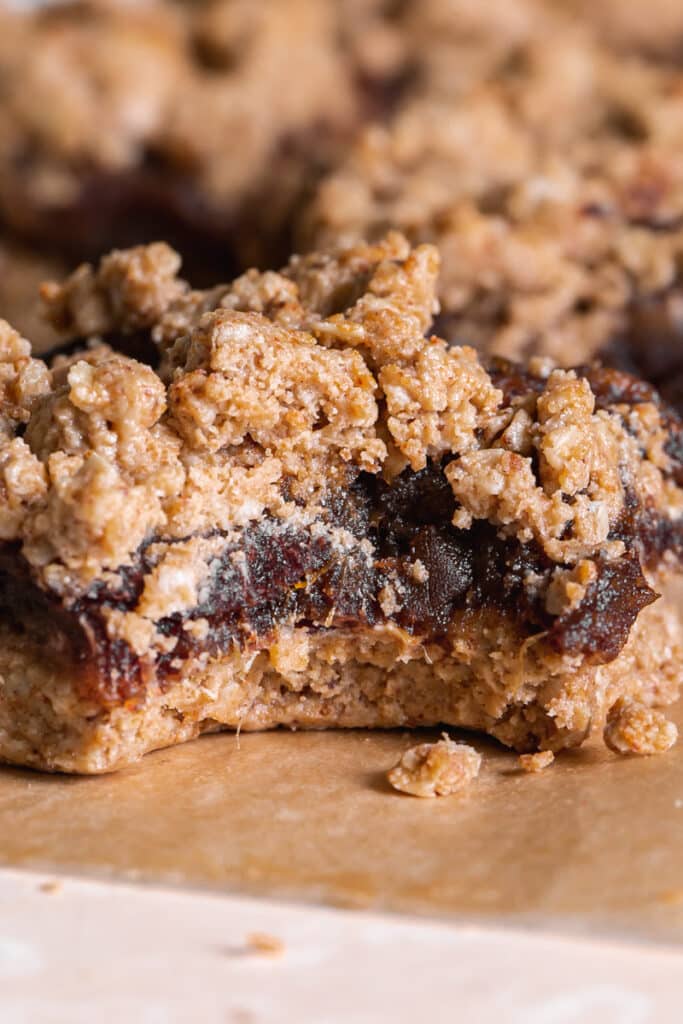 a close up shot of a Peanut Butter Oat Date Crumb Bar with a bite taken out of it on a piece of parchment paper with more bars behind it