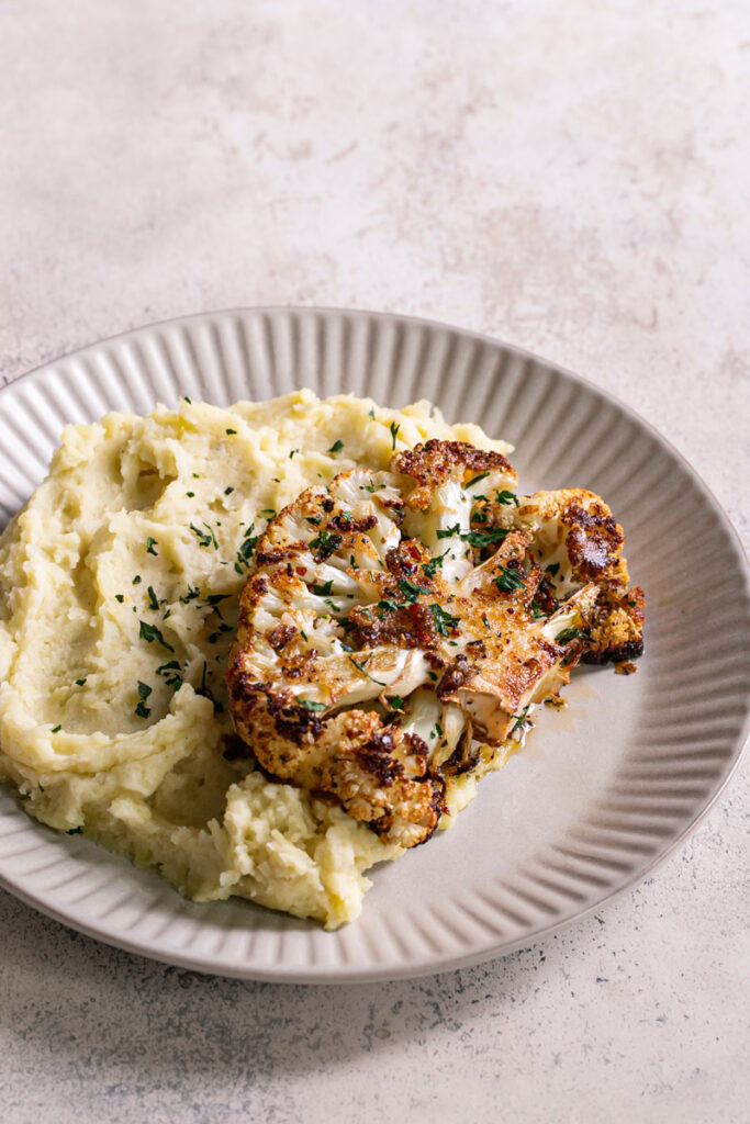 3/4 shot of a dinner plate with mashed potatoes and Kick'n Chick'n Roasted Cauliflower Steak on top garnished with fresh parsley