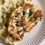 a close up of a dinner plate with mashed potatoes and Kick'n Chick'n Roasted Cauliflower Steak on top garnished with fresh parsley