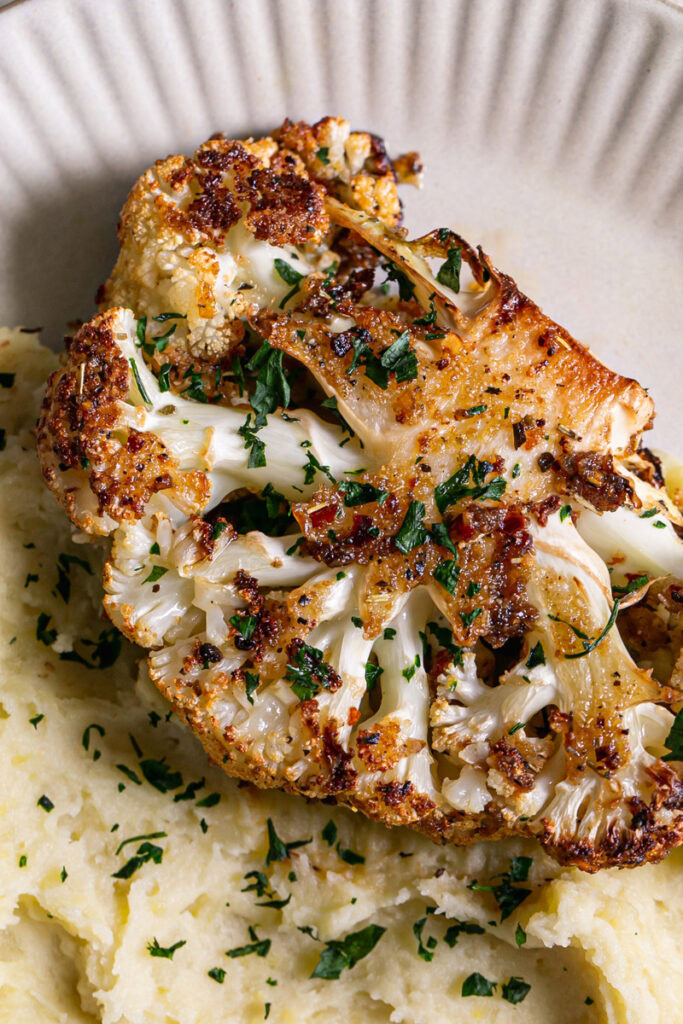 a close up of a dinner plate with mashed potatoes and Kick'n Chick'n Roasted Cauliflower Steak on top garnished with fresh parsley