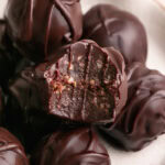 a close up of a plate of chocolate crunch date truffles piled on top of one another, one with a bite taken out of it