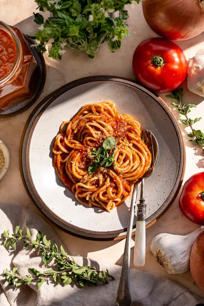 spaghetti noodles tossed in the tomato sauce in a pasta bowl with a spoon and fork surrounded by fresh tomatoes, garlic and oregano