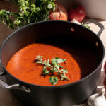 a pot of Caramelized Onion and Garlic Roasted Tomato Sauce with fresh basil, onion, oregano, and tomatoes surrounding it