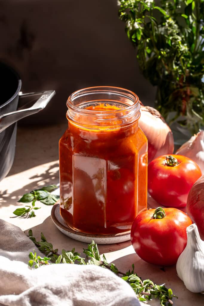 a jar of Caramelized Onion and Garlic Roasted Tomato Sauce surrounded by fresh tomatoes, onion, basil and garlic